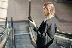 Portrait of woman on escalator, using mobile phone and looking at smartphone screen, standing in black suit on street photo