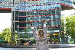 Corporate people. Rear view of businesswoman raising hands up in celebration, triumphing, achieve goal and rejoicing, standing on street photo