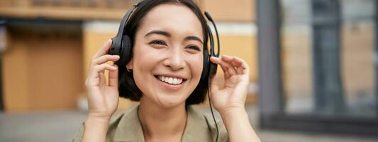 Portrait of beautiful asian woman in headphones, listening music on street of city centre, smiling happily photo