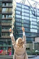 Rear view of corporate woman, lawyer celebrating, lifting hands up and triumphing, achieve goal or success, standing outside on street of city center photo