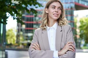 Portrait of young successful businesswoman in suit, cross arms on chest, smile and look confident, standing on street of business center photo
