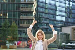 Corporate woman celebrating her victory outside on street. Happy businesswoman raising hands up and triumphing from excitement photo