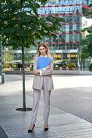 Vertical shot of young businesswoman in beige suit, holding documents in hands, looking confident at camera, standing outdoors in city photo