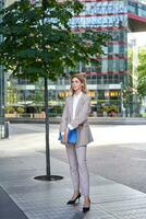 Portrait of young saleswoman in beige business suit, holding blue folder with work documents, standing outdoors on street of city center photo