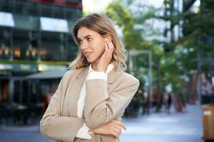 cerca arriba retrato de rubio mujer de negocios, confidente corporativo mujer en auriculares y beige traje, posando al aire libre en calle de ciudad centrar foto