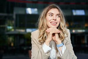 Beautiful corporate woman, looks dreamy and smiles, stands outside on street, leans her head on hands, daydreaming, resting on break after office work photo