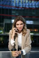 Vertical shot of smiling businesswoman answer mobile phone call, talking on smartphone and drinking coffee photo