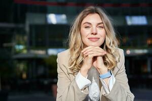 Beautiful corporate woman, looks dreamy and smiles, stands outside on street, leans her head on hands, daydreaming, resting on break after office work photo