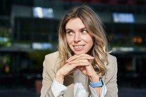 Young smiling business woman in suit, looks confident, stands relaxed on street, poses near office buildings photo