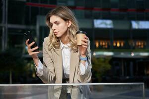 Corporate people. Young businesswoman drinking coffee, using smartphone app, resting on lunch break photo