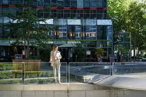 Corporate businesswoman in beige suit. Silhouette of young corporate woman with documents and laptop, posing outdoors in city center photo