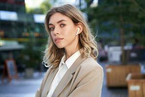 Confident businesswoman in headphones and beige suit, looking at camera with thoughtful gaze, posing outdoors photo