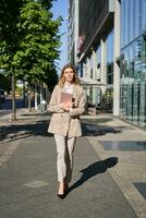 Vertical shot of businesswoman walking on street with digital tablet, going to work, wearing beige suit and high heels photo