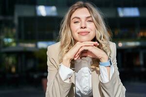 Beautiful corporate woman, looks dreamy and smiles, stands outside on street, leans her head on hands, daydreaming, resting on break after office work photo