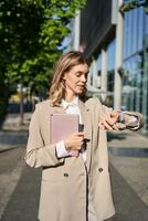 Vertical portrait of beautiful businesswoman in suit, holds her digital tablet, looks at watch on wrist, reads message or checks time photo