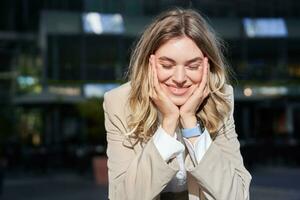 Portrait of beautiful happy corporate woman in suit, stands on street and smiles, poses near office buildings photo