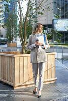 Beautiful businesswoman in suit, drinking takeaway coffee on street, waiting for someone near office building on lunch break photo