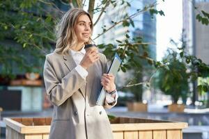 Smiling corporate woman in suit, drinking takeaway coffee on street near office building photo