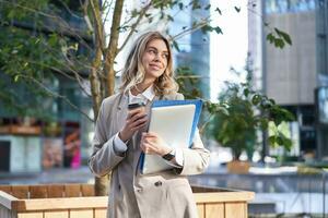 Smiling businesswoman standing on street, holding work documents, laptop and drinking takeaway coffee photo
