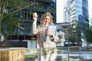 Enthusiastic businesswoman feels upbeat after morning cup of coffee, raise her cup up and dances outside photo