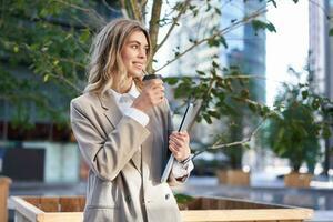 Smiling corporate woman in suit, drinking takeaway coffee on street near office building photo