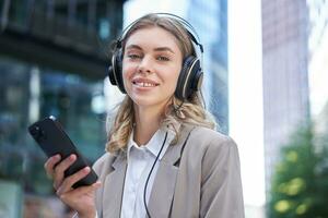 Portrait of smiling young businesswoman listening music in headphones and using mobile phone while on a busy city street photo