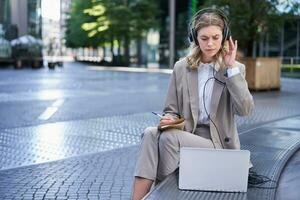 Businesswoman looking perplexed at laptop, taking notes in her notebook, attending online work meeting while sitting on street photo