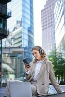 corporativo mujer en traje, sentado en ciudad, escuchando a música con computadora portátil, desplazamiento Noticias en su móvil teléfono, relajante en almuerzo descanso foto