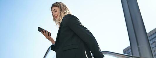 Rear view of young business woman in black blazer, looking at smartphone screen and smiling, standing outdoors on escalator photo