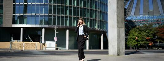 Young corporate woman in suit walking on street and talking on mobile phone. Businesswoman having telephone conversation while going to work photo