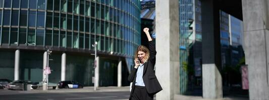 Excited businesswoman receives great news, celebrating while talking on mobile phone. Corporate woman shouts from joy on street, triumphing photo