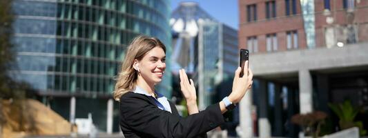 Portrait of businesswoman wave her hand at mobile phone camera, waves hand during video chat, stands in suit in city center outdoors photo