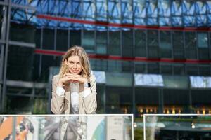 Beautiful young woman in beige suit, standing near office buildings in city center, smiling and looking dreamy photo