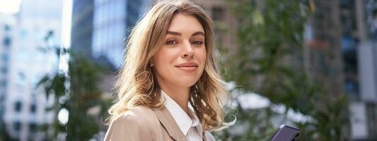 Close up portrait of corporate woman, young intern walks on street to office, holds mobile phone, texts messages on smartphone photo