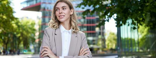 Portrait of young successful businesswoman in suit, cross arms on chest, smile and look confident, standing on street of business center photo