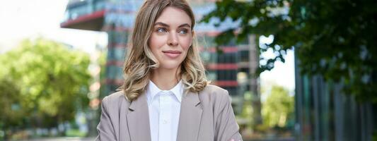 Portrait of young successful businesswoman in suit, cross arms on chest, smile and look confident, standing on street of business center photo