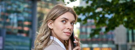 Close up shot of businesswoman talking on mobile phone. Corporate woman calling someone, looking around, standing outdoors photo