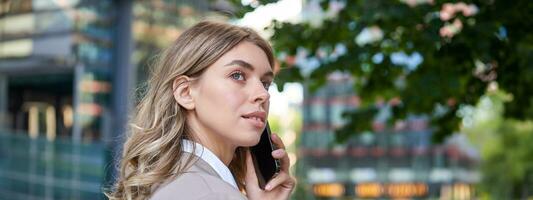 Close up shot of businesswoman talking on mobile phone. Corporate woman calling someone, looking around, standing outdoors photo