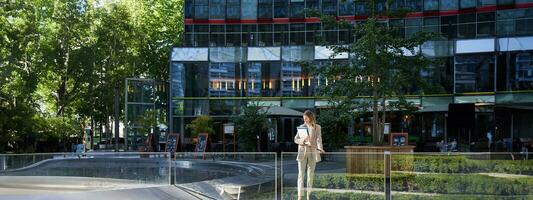 Corporate businesswoman in beige suit. Silhouette of young corporate woman with documents and laptop, posing outdoors in city center photo