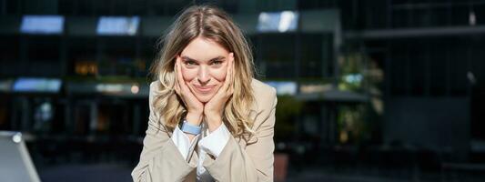 Portrait of beautiful happy corporate woman in suit, stands on street and smiles, poses near office buildings photo