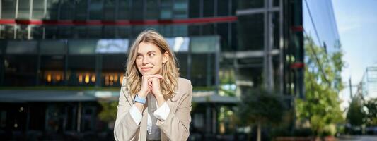 Beautiful young woman in beige suit, standing near office buildings in city center, smiling and looking dreamy photo