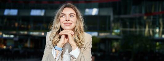 Beautiful corporate woman, looks dreamy and smiles, stands outside on street, leans her head on hands, daydreaming, resting on break after office work photo