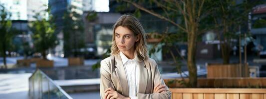 cerca arriba retrato de confidente y exitoso mujer de negocios en traje, cruzar brazos en pecho, en pie en poder actitud en calle cerca oficina edificio foto