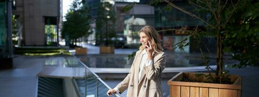 Beautiful young businesswoman in suit talks on mobile phone on street. Corporate woman calls someone on phone while stands outside photo