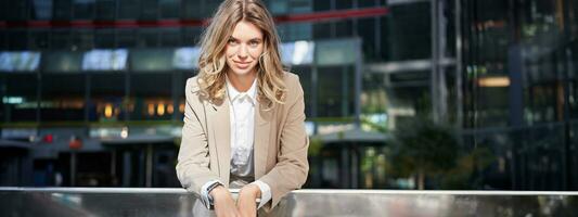 Young beautiful businesswoman standing in beige suit on sunny street near office buildings photo