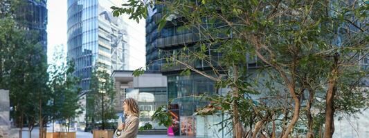 Vertical shot of corporate woman in beige suit standing outside on street, drinks morning coffee takeaway before work photo