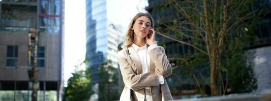 Confident corporate woman looks at camera stands on street. Businesswoman in beige suit, posing in city near office buildings photo