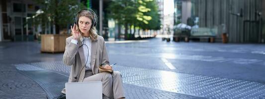 Woman sitting on a street with laptop and headphones plugged in, taking notes. Corporate worker attend online team meeting and writing down information, working outdoors photo