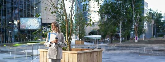 Beautiful businesswoman in suit, drinking takeaway coffee on street, waiting for someone near office building on lunch break photo