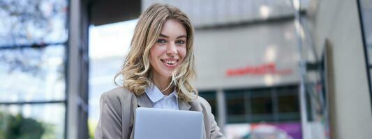 retrato de joven corporativo mujer, participación su ordenador portátil y sonriente, yendo a trabajar, vistiendo profesional atuendo, beige suite foto